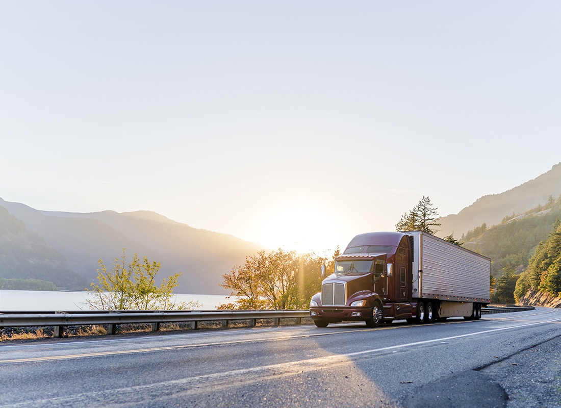 Insurance Solutions - View of a Red Tractor Railer Driving on an Empty Highway at Sunset with Scenic Views of a River and Mountains in the Background